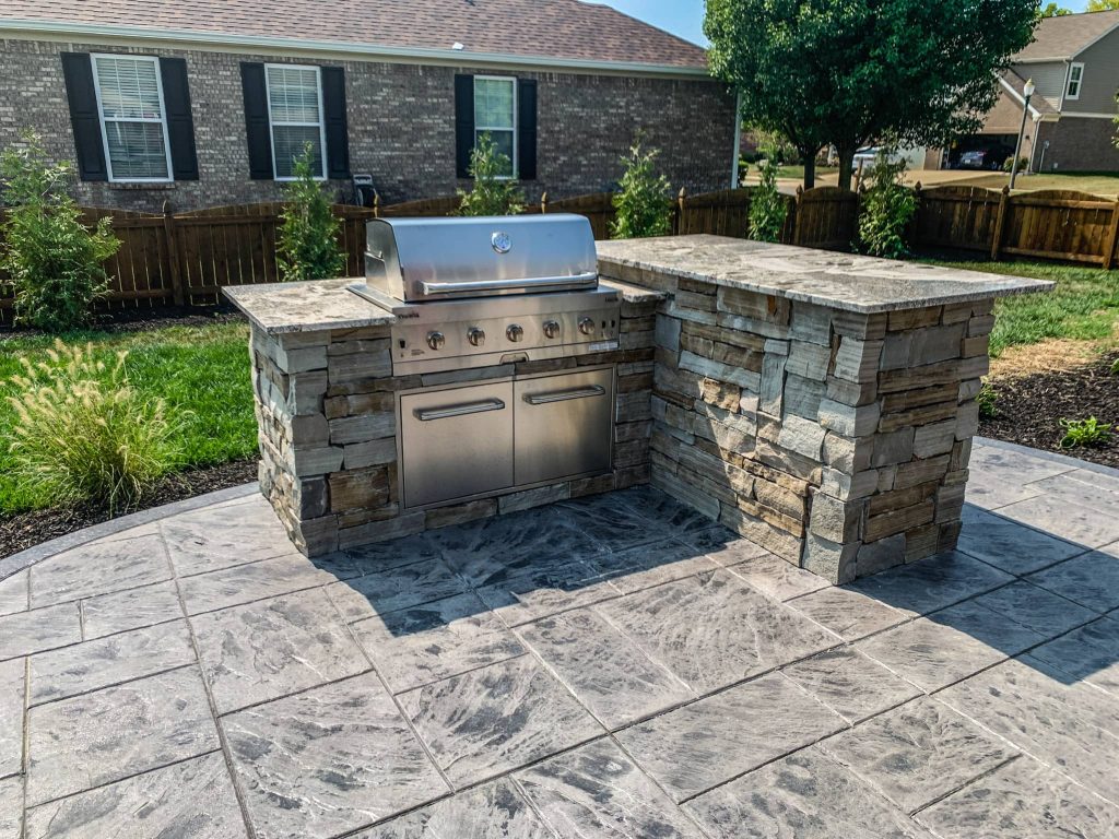 Custom-built concrete patio with an outdoor kitchen and stainless steel grill, set against a brick house backdrop.