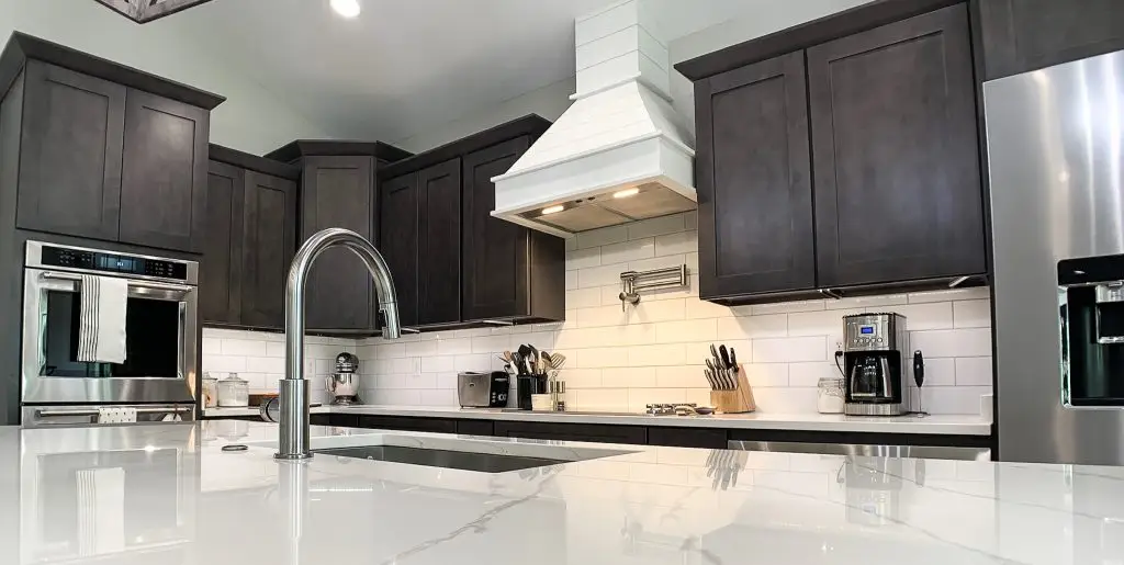 Modern kitchen featuring dark shaker-style cabinets, a built-in stainless steel oven, white subway tile backsplash, and a sleek white quartz island with a gooseneck faucet.