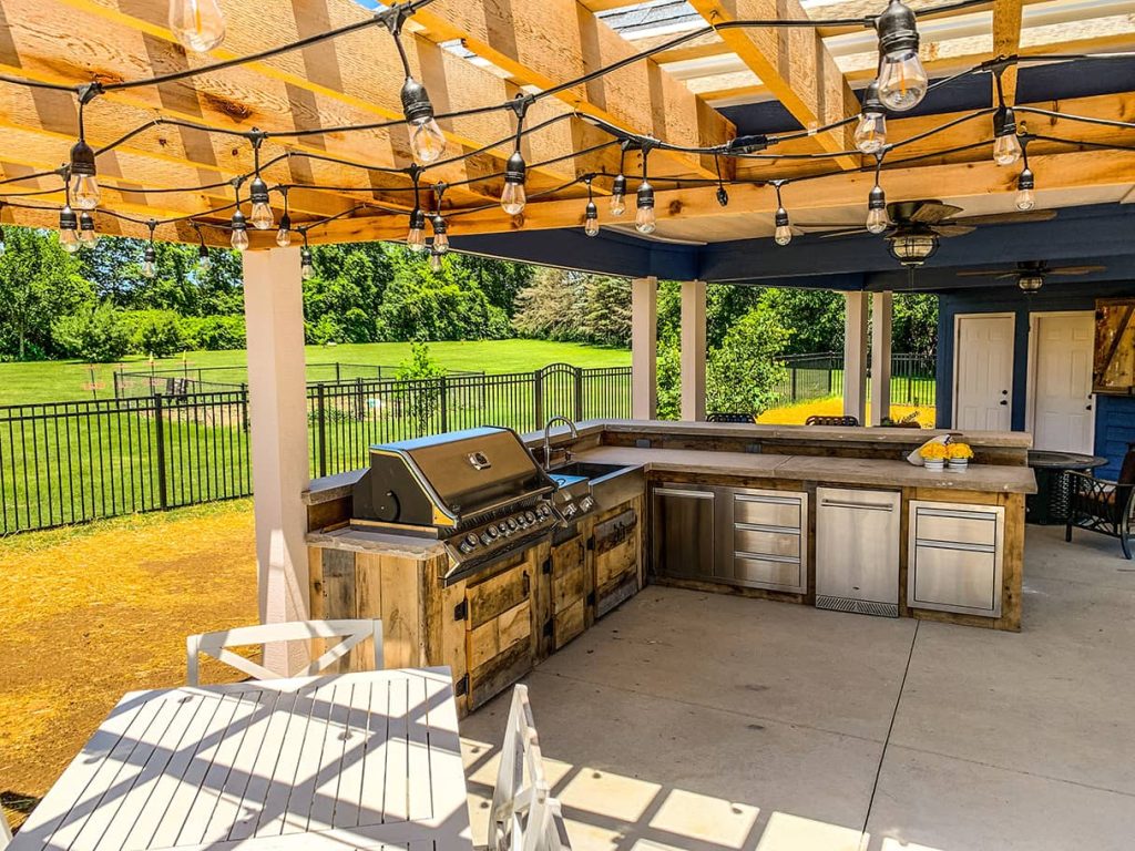 Covered outdoor kitchen with rustic wood cabinetry, stainless steel appliances, and exposed string lights under a wooden pergola roof