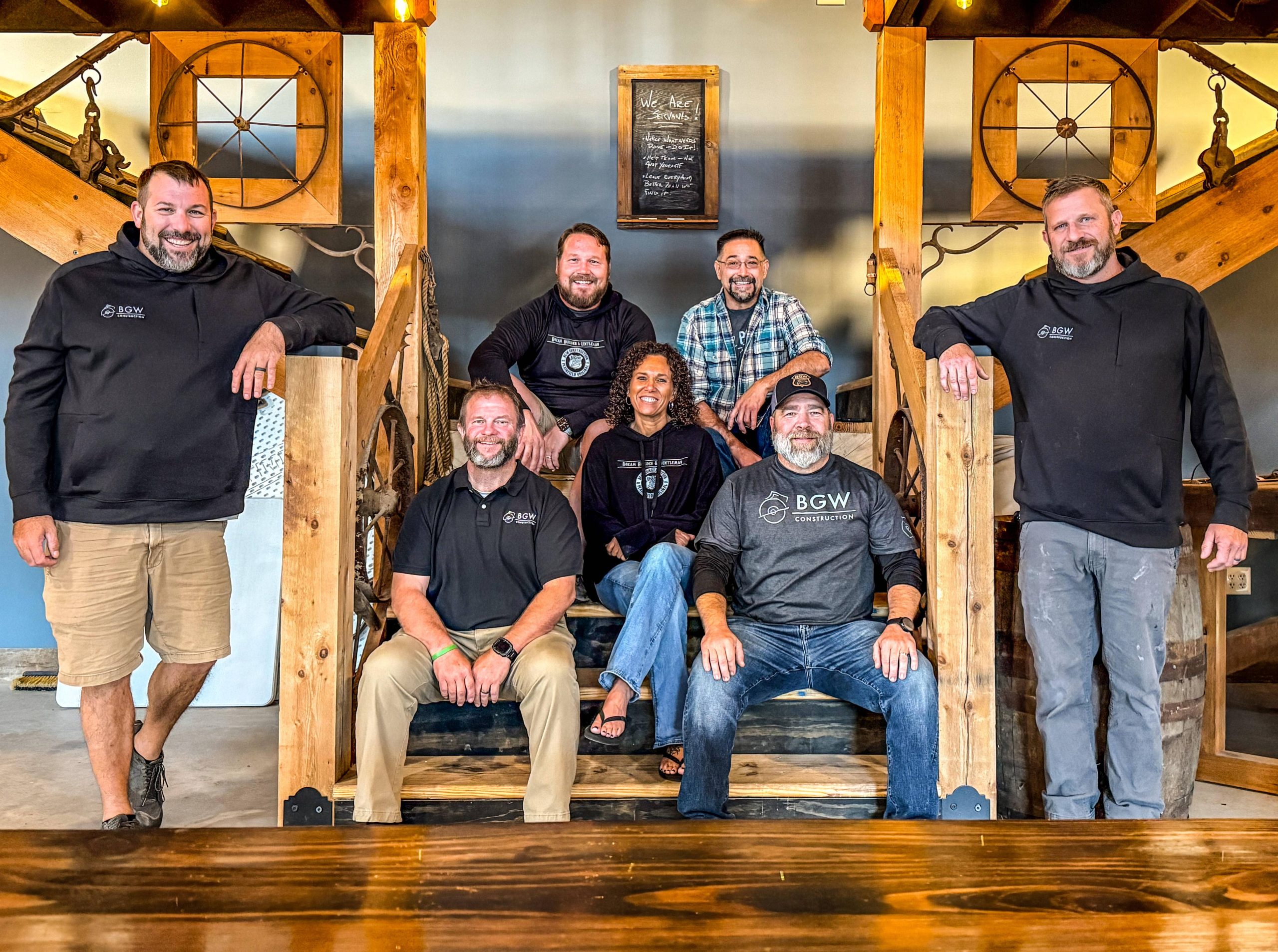 Group photo of BGW Construction LLC team members sitting and standing on wooden stairs in a rustic office setting, all smiling and wearing branded apparel.