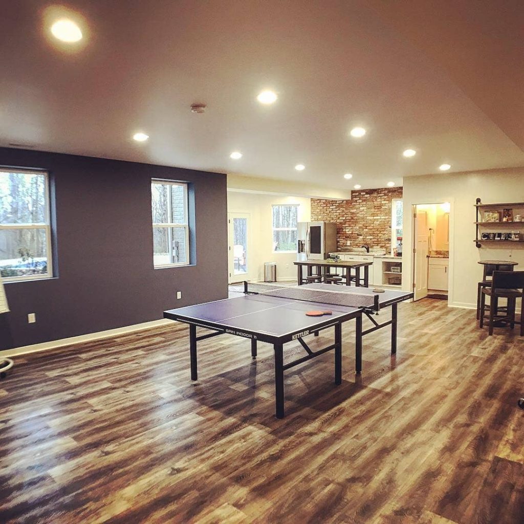 Modern game room with a ping pong table in the center, featuring wood-look flooring, recessed lighting, and an open-concept layout with kitchen and bar area.