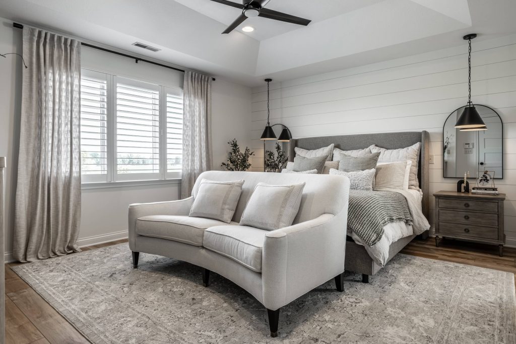 Modern neutral-toned bedroom with a gray upholstered bed, layered bedding, white curved loveseat, and soft natural light from shuttered windows.