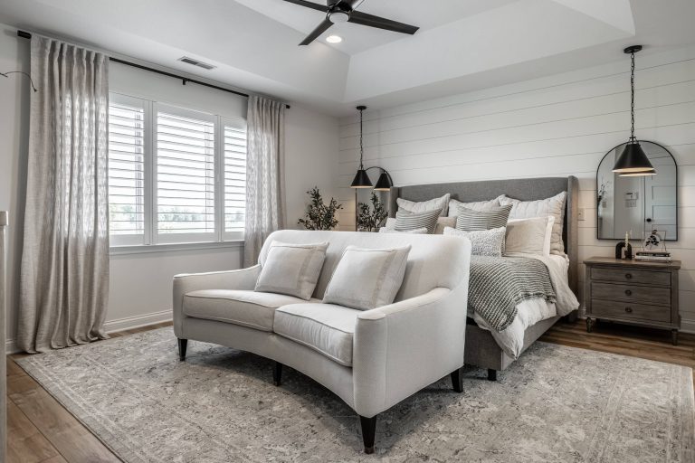Modern neutral-toned bedroom with a gray upholstered bed, layered bedding, white curved loveseat, and soft natural light from shuttered windows.