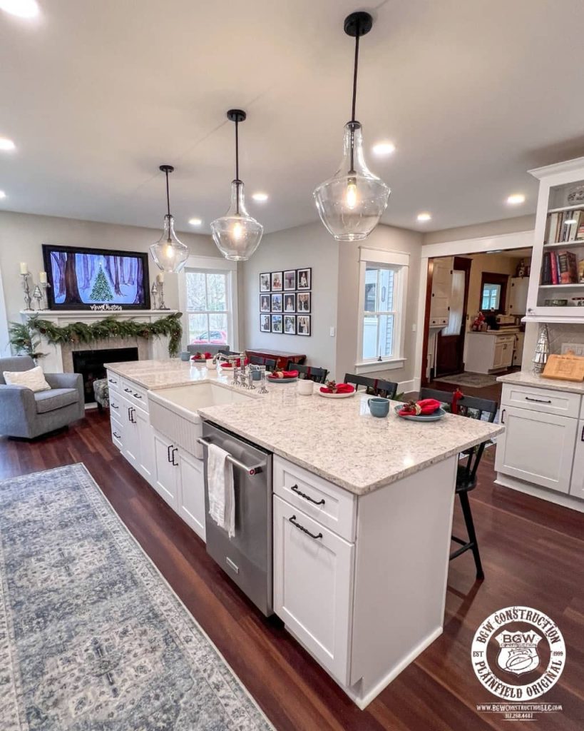 Open-concept kitchen remodel featuring white shaker cabinets, large quartz island with seating, farmhouse sink, pendant lighting, and view into living area with holiday decor