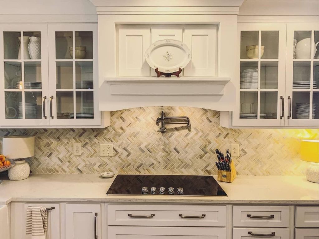 White kitchen with glass-front cabinets, herringbone tile backsplash, and built-in cooktop under a decorative range hood.