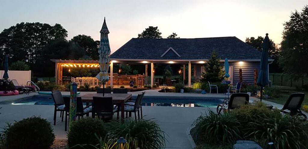 Backyard renovation in Avon, Indiana featuring a swimming pool, covered patio with lighting, pergola seating area, and outdoor dining space at dusk.