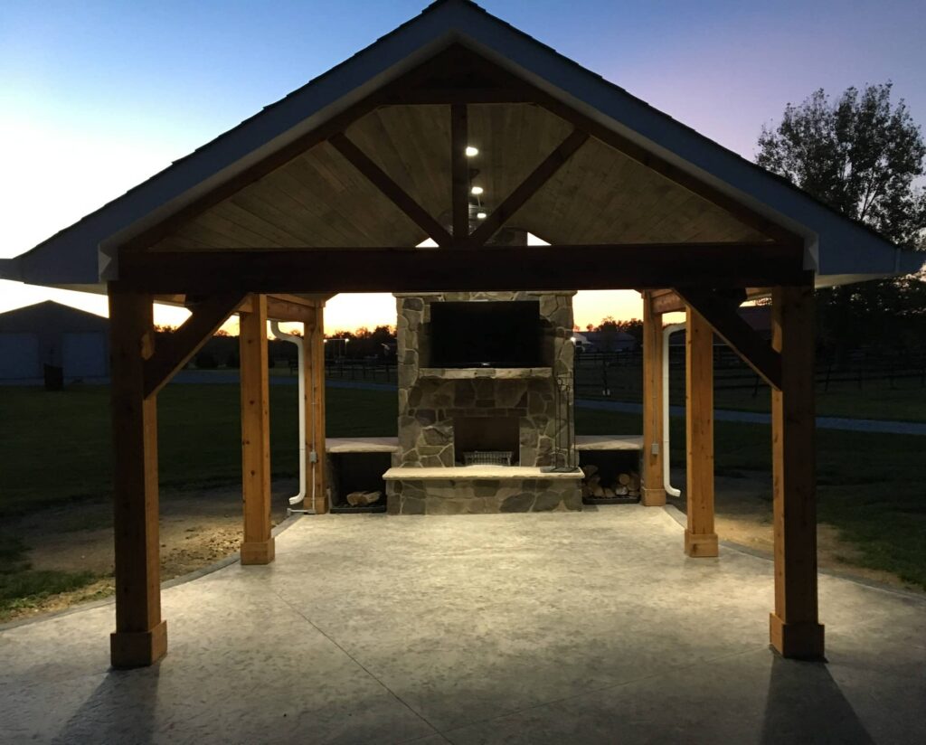 Outdoor pavilion with wooden beams and gable roof featuring a stone fireplace and mounted TV at sunset.