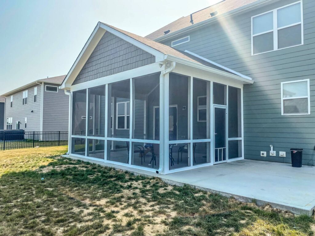 Screened-in backyard porch with white framing attached to a gray suburban house, featuring a covered patio and outdoor seating.