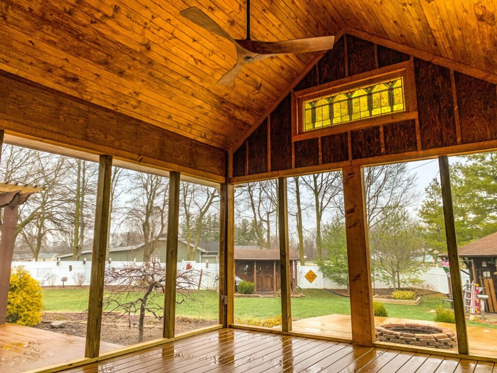 Screened-in wooden porch with vaulted ceiling, ceiling fan, and stained glass window overlooking a backyard with lawn, trees, and fire pit.