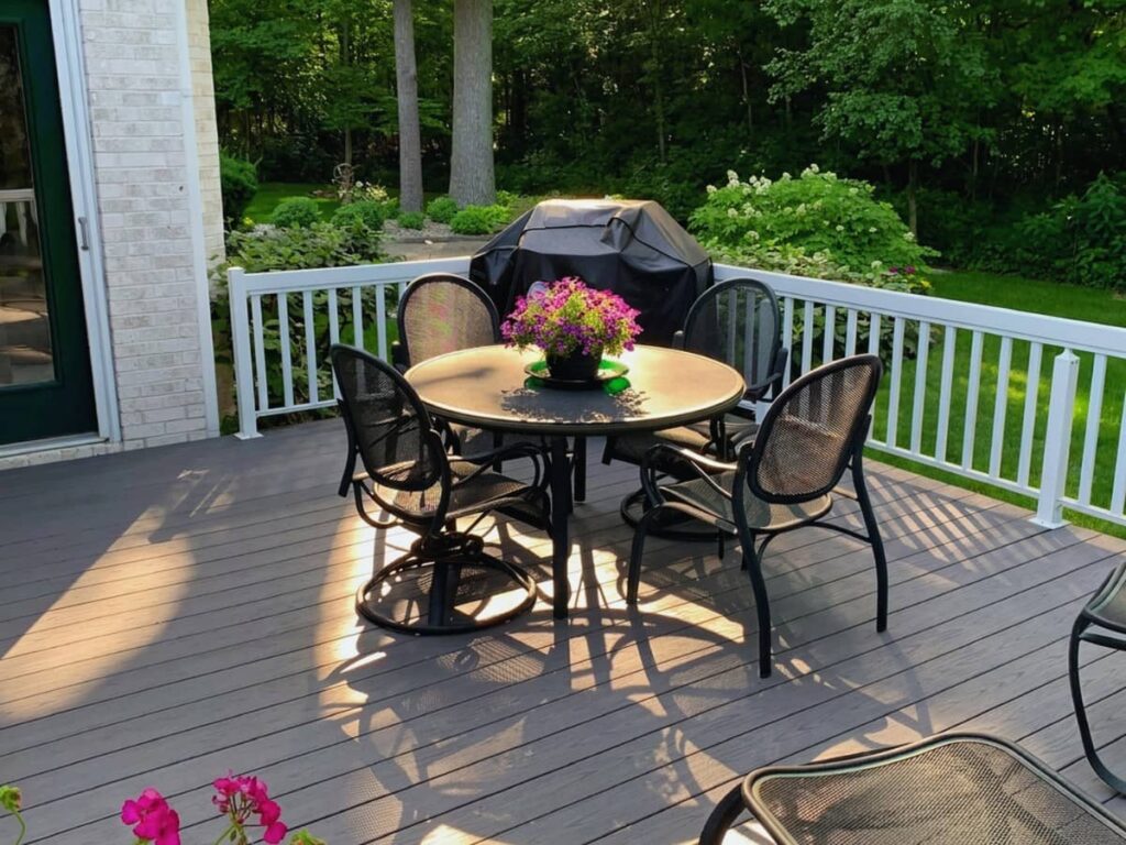 Composite backyard deck with white railing, outdoor dining table, grill, and landscaped wooded backyard.