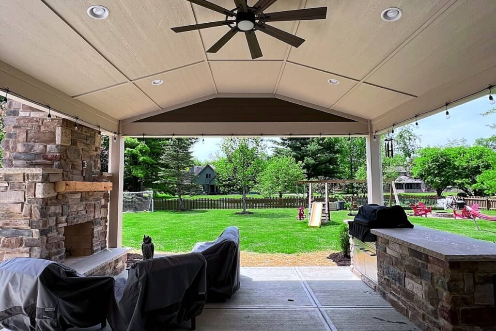 Covered outdoor kitchen with a built-in grill, stone fireplace, vaulted ceiling, and backyard entertainment space.