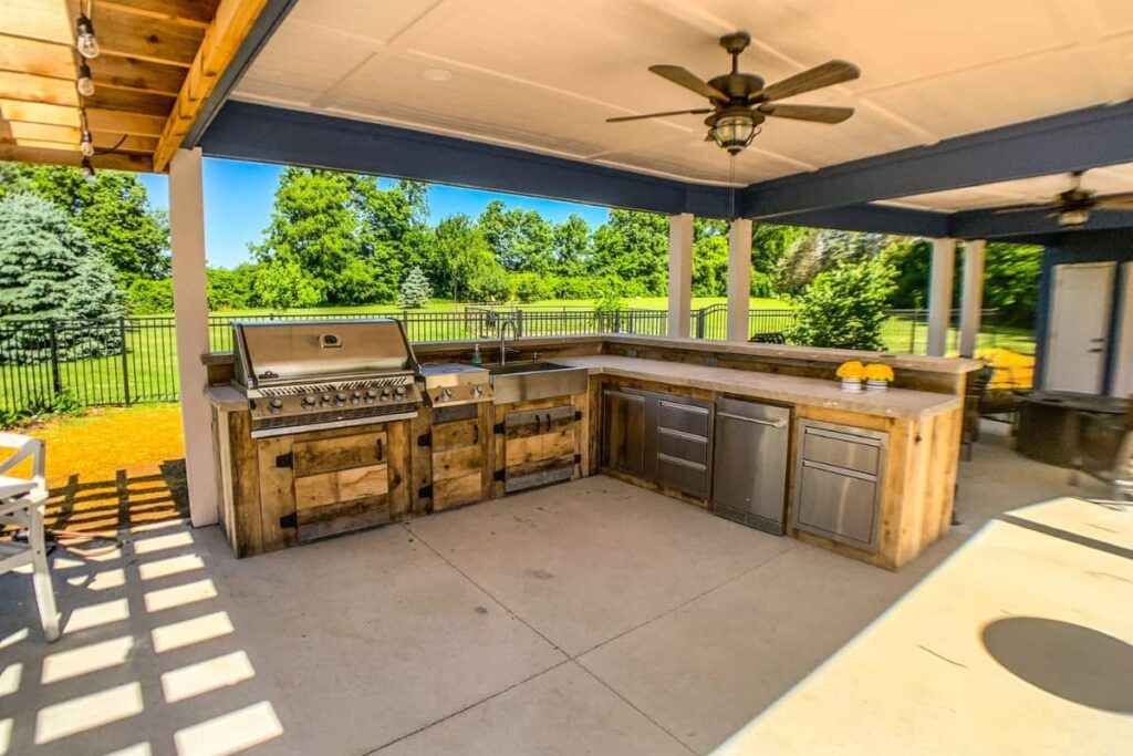 Covered outdoor kitchen with rustic wood cabinetry, built-in grill, sink, refrigerator, and spacious countertop for outdoor entertaining.