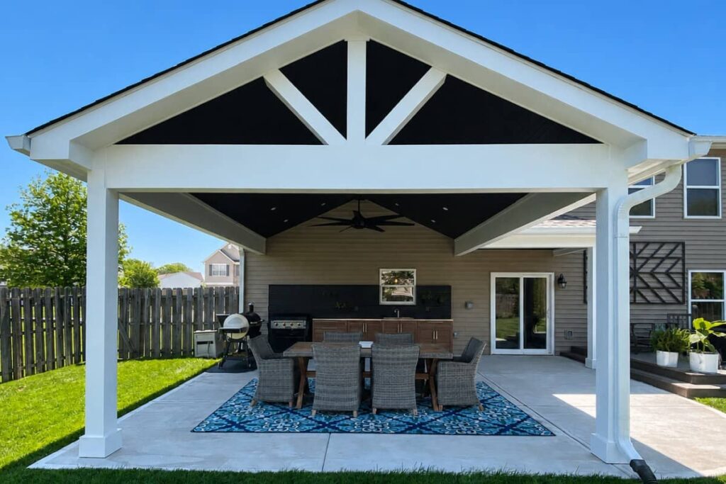 Covered backyard patio with vaulted gable roof, outdoor kitchen, dining table, and seating area beneath a modern timber pavilion.