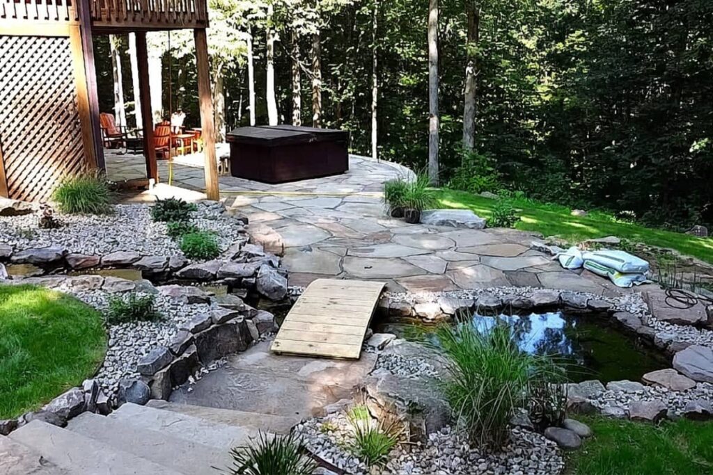 Natural stone patio with a hot tub, decorative pond, small footbridge, and landscaped backyard surrounded by mature trees.