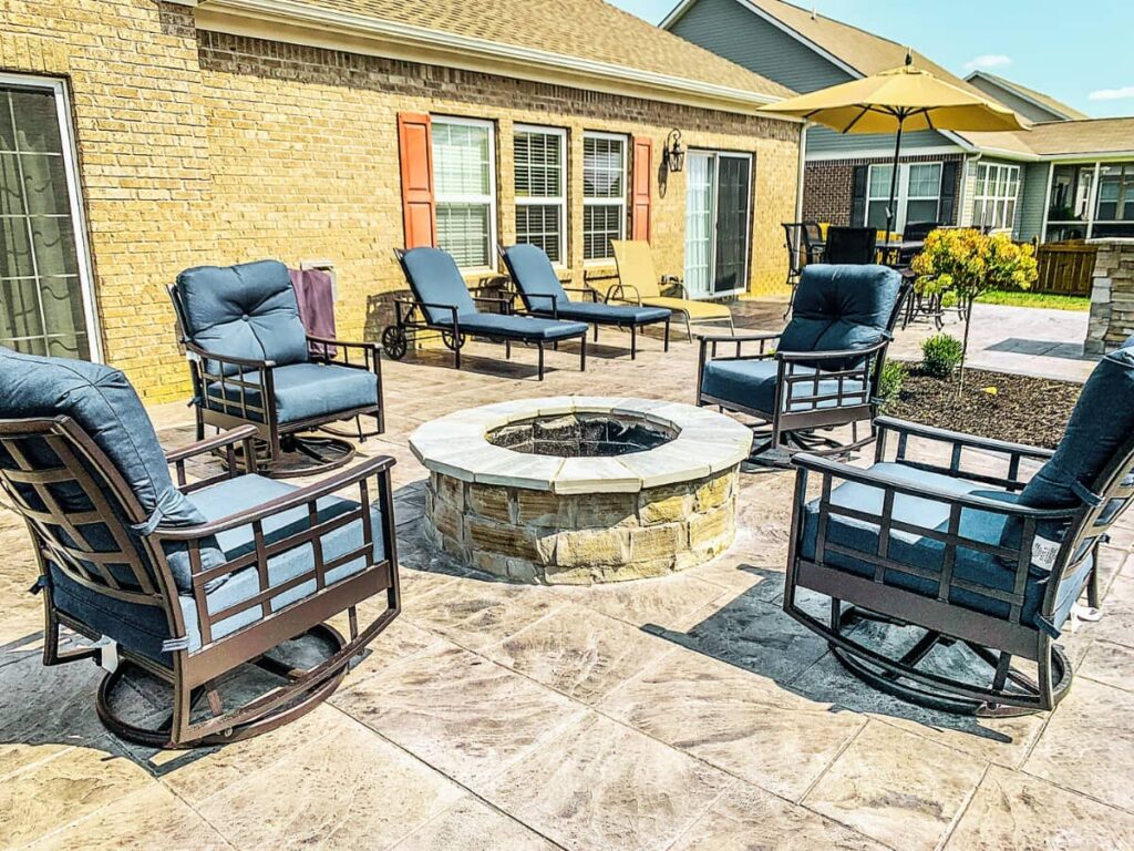 Stamped concrete backyard patio with a circular stone fire pit, cushioned patio chairs, lounge chairs, and umbrella seating outside a brick home.