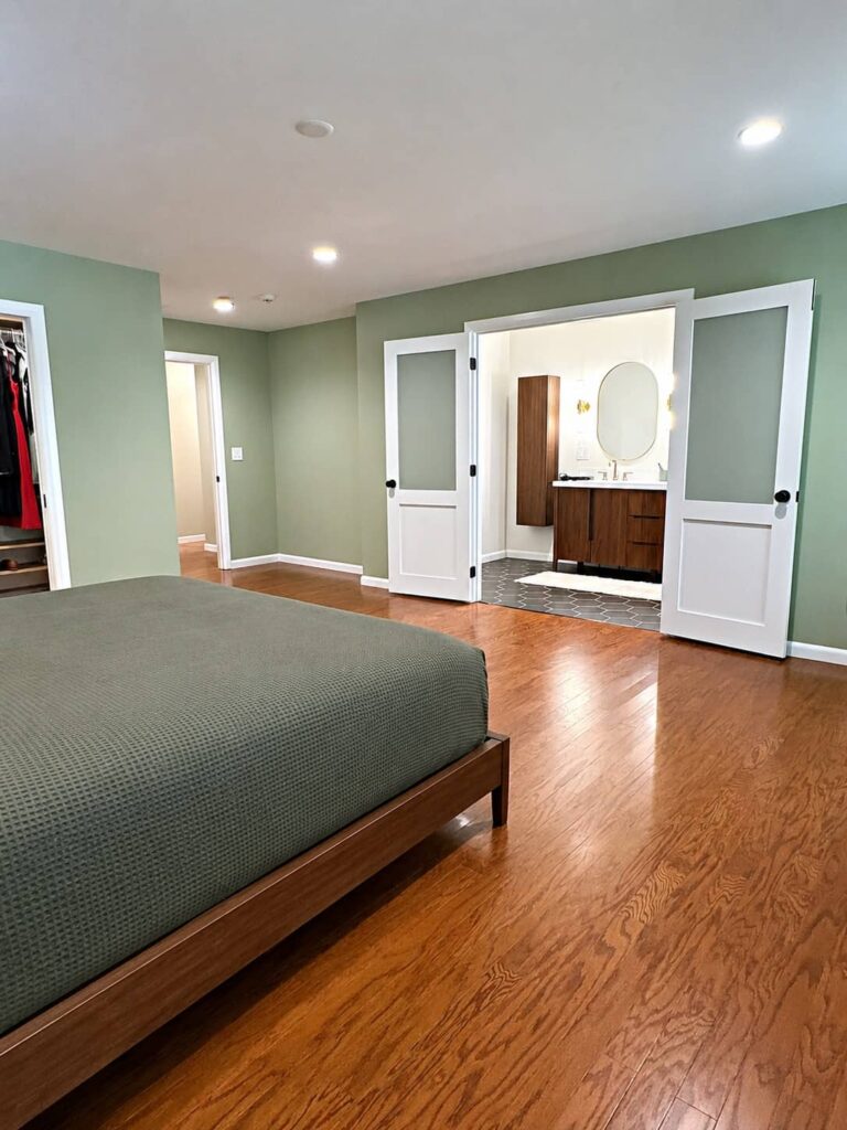 Modern primary bedroom with hardwood flooring, sage green walls, and double doors opening to an ensuite bathroom featuring a walnut vanity and contemporary finishes.
