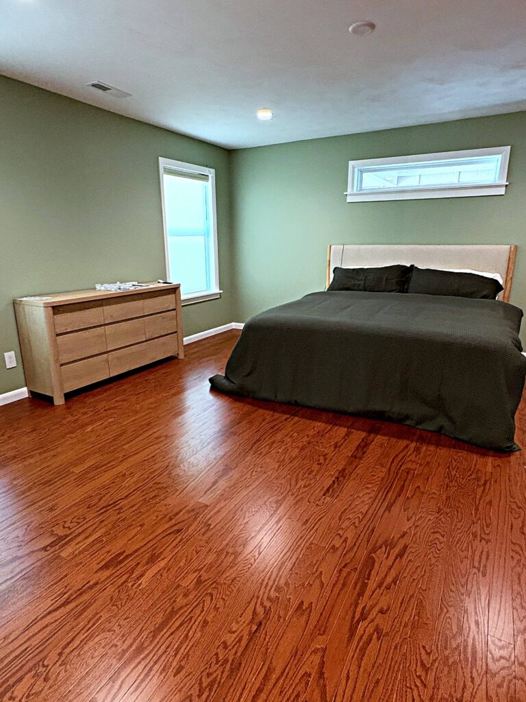 Spacious modern primary bedroom with warm hardwood flooring, sage green walls, upholstered bed, contemporary dresser, and large windows providing natural light.