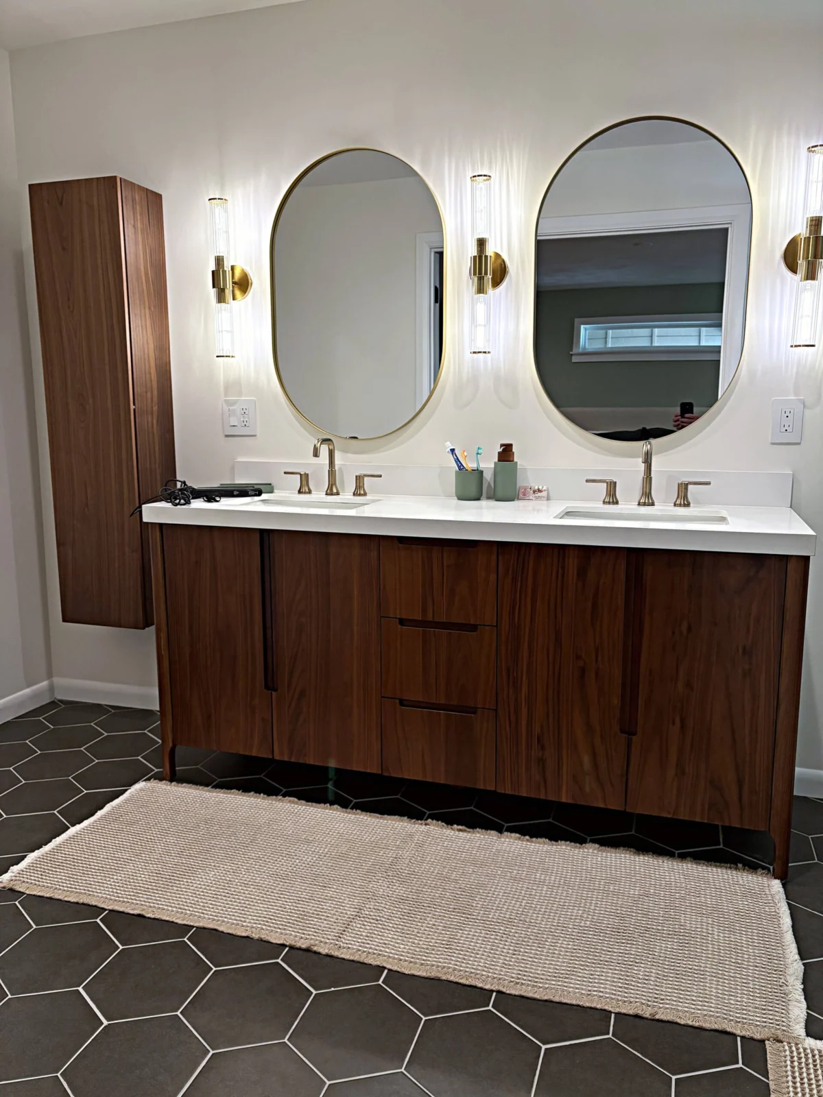 Wood double vanity with a white counter, two oval mirrors, brass-colored fixtures, and dark hexagonal floor tile.