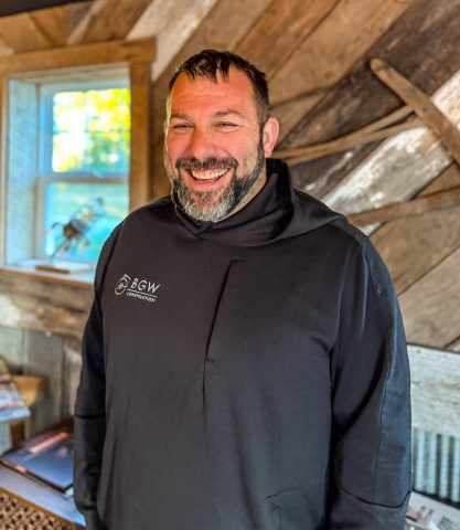 Portrait of Kevin Ladd, smiling and wearing a black BGW Construction hoodie, standing in a rustic, wood-accented room.