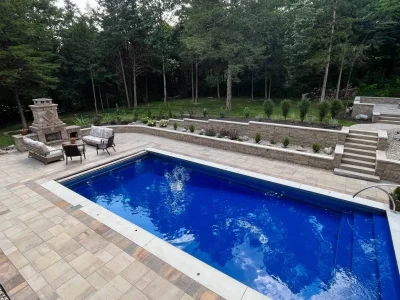 Outdoor Pool and Living Space by BGW Outdoor Living A luxurious backyard featuring a blue in-ground pool, a stone fireplace with seating, and elegant landscaping against a wooded backdrop.