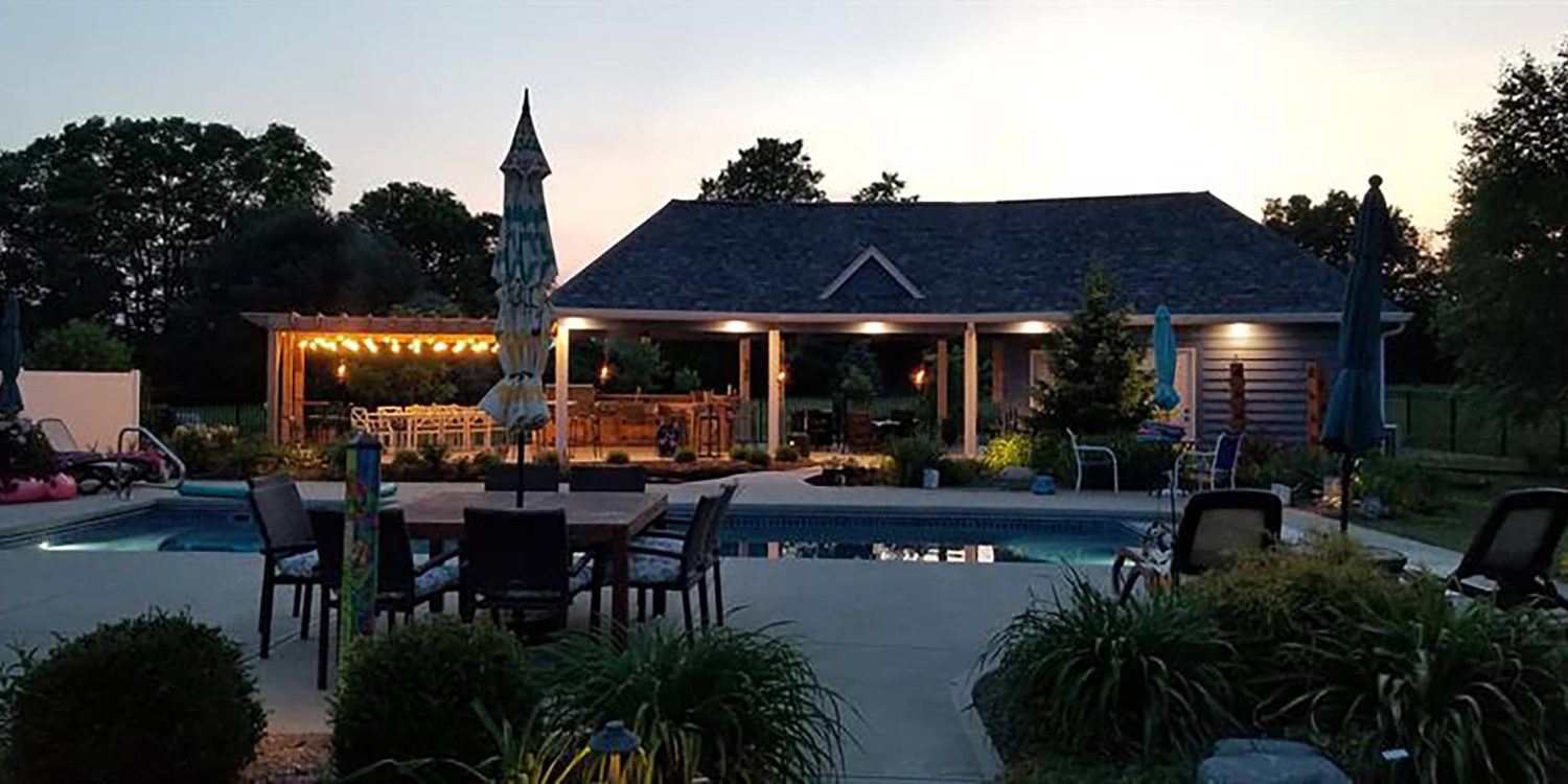 Backyard renovation in Avon, Indiana featuring a swimming pool, covered patio with lighting, pergola seating area, and outdoor dining space at dusk.