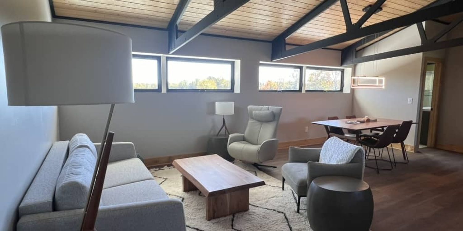 Renovated barn loft living area with light gray sofa, modern armchairs, wood coffee table, and exposed black ceiling beams.