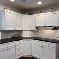 Remodeled kitchen corner with white shaker cabinets, black countertops, and tile backsplash under recessed lighting