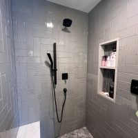 Walk-in shower with gray herringbone tiles, black fixtures, and built-in shelving niche
