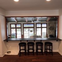 Open concept breakfast bar with black counter and four stools overlooking living room with windows and ceiling beams