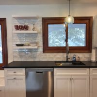 Kitchen sink area with floating white shelves holding glassware and dishes against tile backsplash