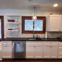 Kitchen sink area with stainless steel dishwasher, black countertops, white cabinets, and window with wood trim