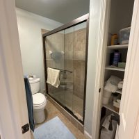 Lake house bathroom before renovation showing a glass-door shower with beige tile, a toilet, and a linen closet with open shelving.