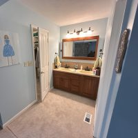 Lake house bathroom before renovation showing double-sink vanity with wood cabinets, large mirror, and soft blue walls.