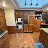 Lake house kitchen before renovation, featuring wood-paneled built-in fridge, double oven, granite countertops, and partial view into rustic living room.