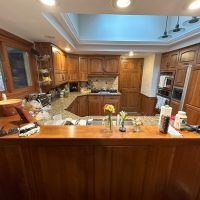 View of a lake house kitchen before renovation, with wooden cabinetry, granite countertops, a central sink, and visible clutter on the counter.