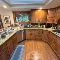 Lake house kitchen before renovation, featuring double sink, granite countertops, wooden cabinetry, and corner prep area under large windows.