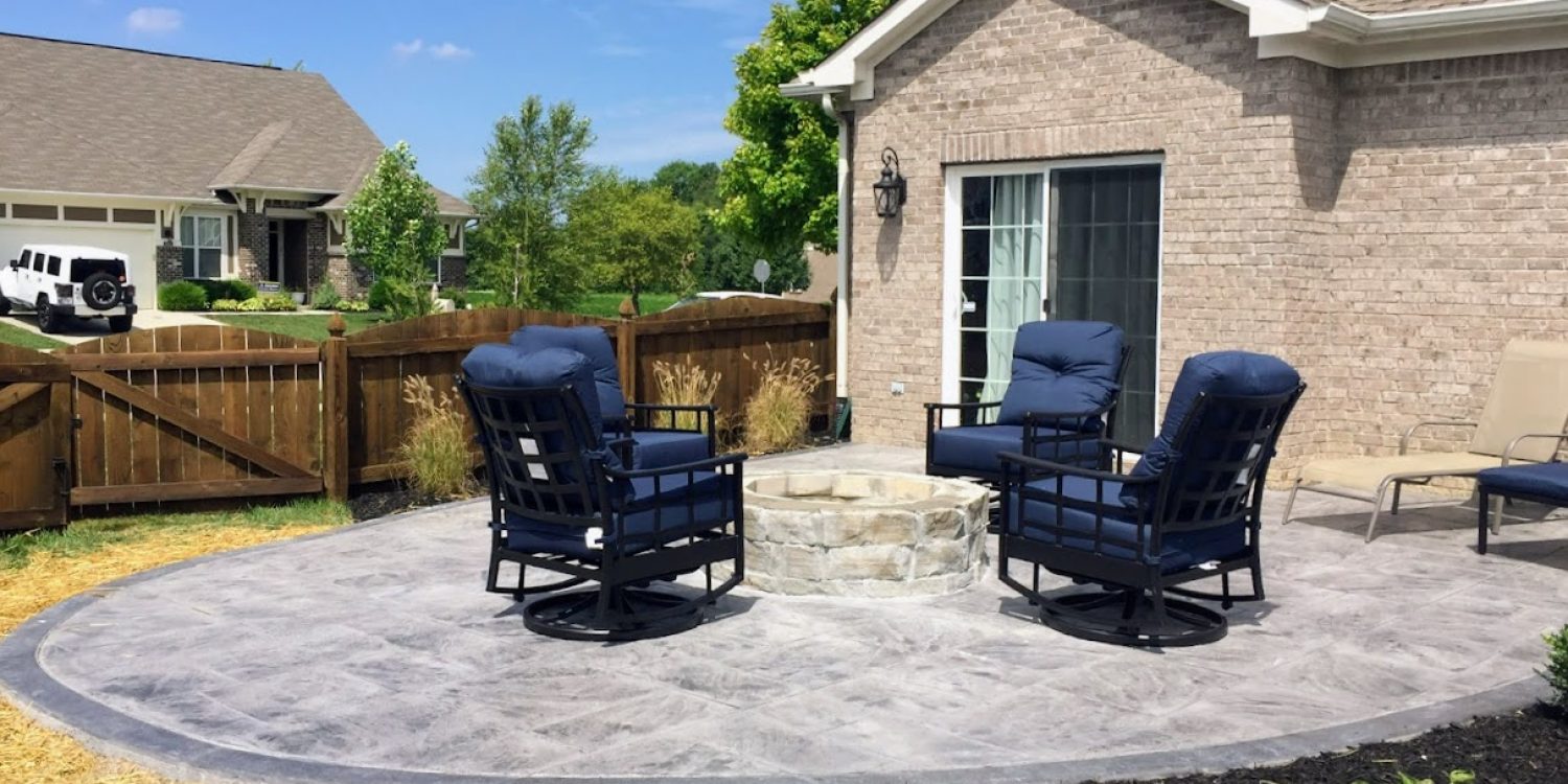 Stamped concrete patio with circular stone fire pit, navy blue cushioned chairs, and minimal landscaping in a fenced suburban backyard.