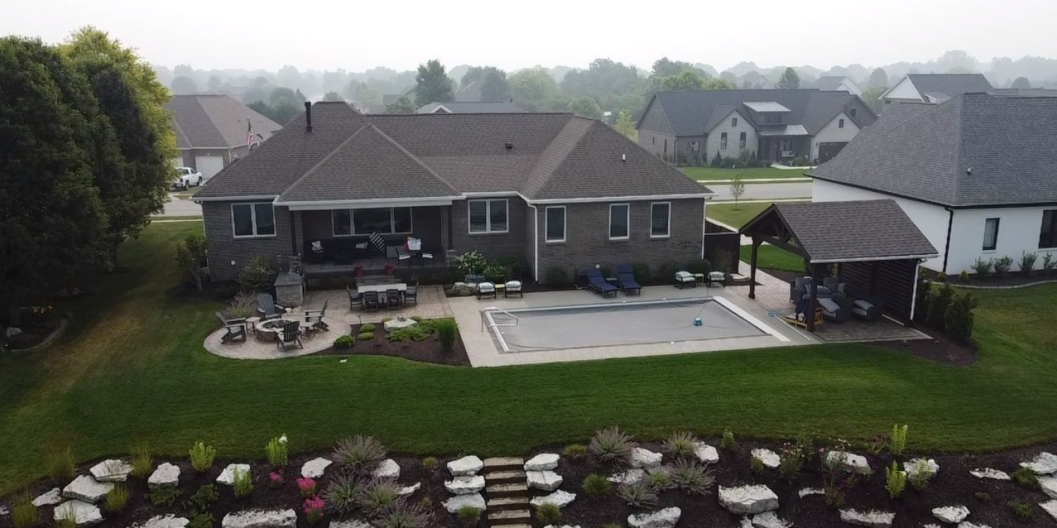 Aerial view of a luxury home with a landscaped backyard featuring a swimming pool, patio seating, a fire pit area, and a covered outdoor lounge.