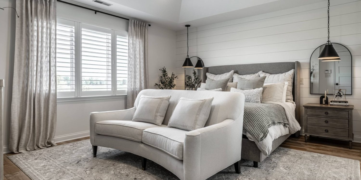 Modern neutral-toned bedroom with a gray upholstered bed, layered bedding, white curved loveseat, and soft natural light from shuttered windows.