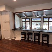 Wide kitchen bar with black countertop and four dark stools facing open living room with large windows