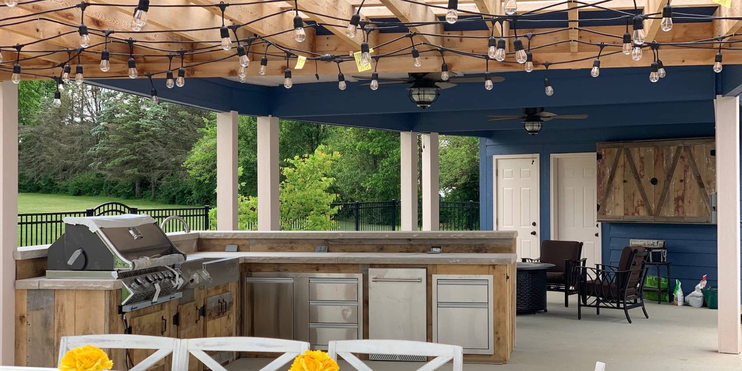 Covered outdoor kitchen with pergola, string lights, stainless steel grill, and bar seating in Plainfield, Indiana.