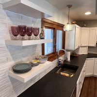 Floating white shelves displaying dishes and glassware against a textured white kitchen backsplash
