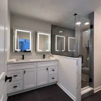 White double sink vanity beside a glass shower with LED mirrors and black fixtures